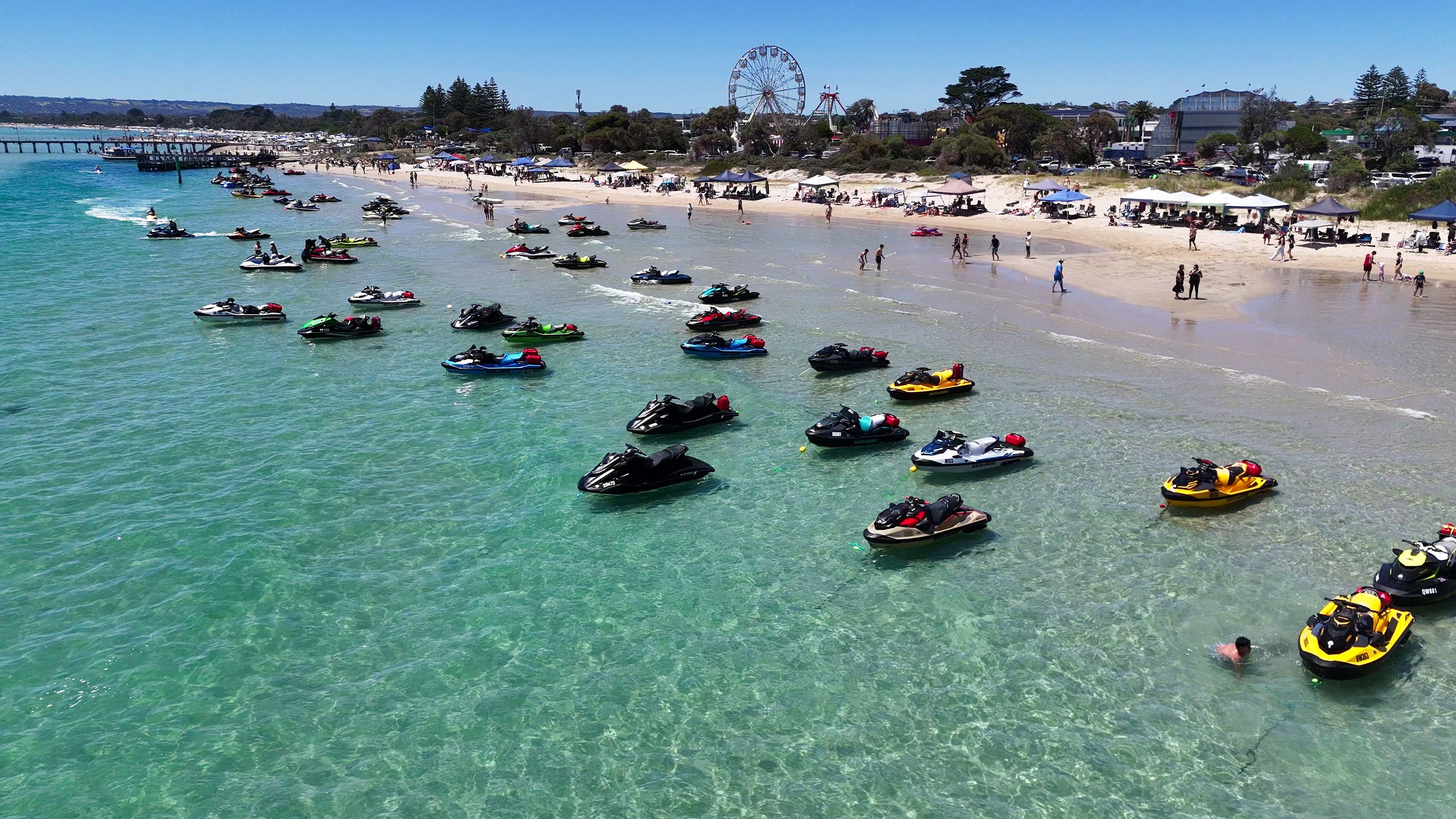 Aerial view of dozens of jet skis floating in turquoise water at a packed beach with ferris wheel in background